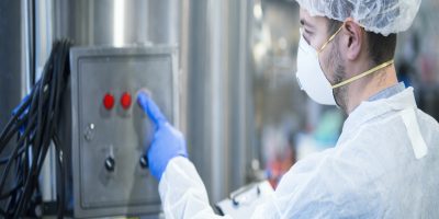 Technologist in white protective uniform with hairnet and mask operating on industrial machine for food processing.